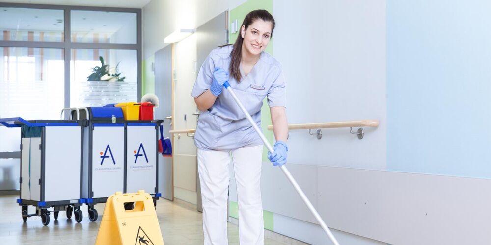 Mitarbeiterin wischt den Boden in einem Krankenhausflur. Neben ihr steht ein Hinweisschild, dass der Boden rutschig ist. Außerdem steht hinter ihr ein Wagen mit Putzutensilien.  