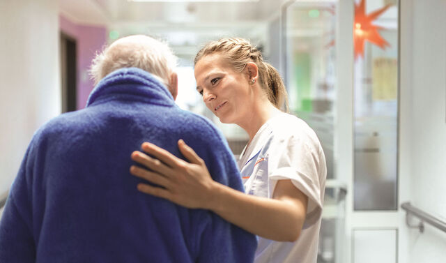 Eine Krankenpflegerin legt einem Senioren eine Hand auf den Rücken und begleitet diesen durch den Krankenhausflur.