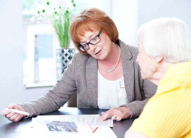 Eine Frau sitzt mit einer Seniorin an einem Tisch und berät diese. 