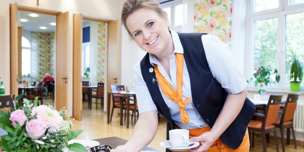 Eine Servicemitarbeiterin nimmt einen Teller von einem Tisch und hat eine Tasse in der Hand. Sie schaut dabei lächelnd in die Kamera.