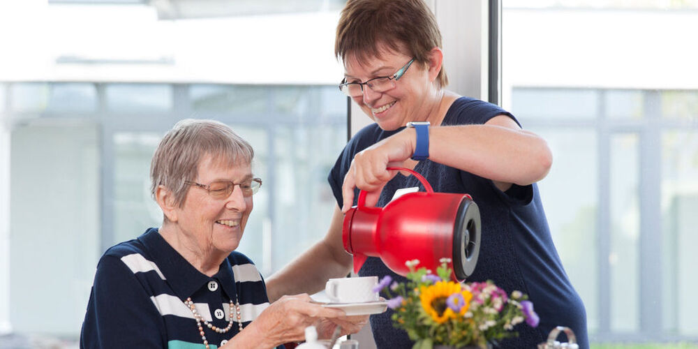 Das Bild zeigt eine Frau, die einer älteren Dame einen Kaffee einschenkt. 