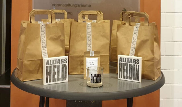 Drei Papiertüten liegen auf einem runden Tisch. Davor sind zwei Postkarten aufgestellt mit den Wörtern "Alltagsheld" und "Alltagsheldin".