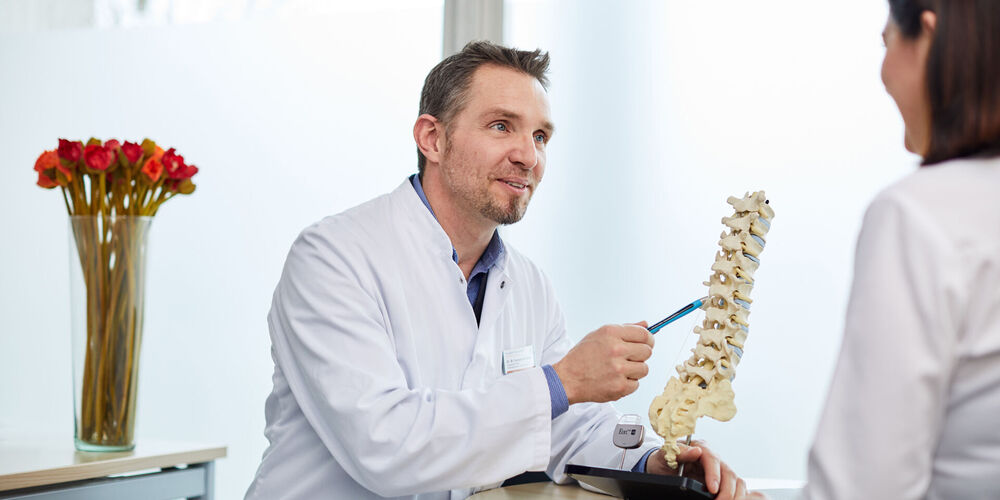 Fachbereich Wirbelsäulentherapie Das Bild zeigt einen Arzt, der einer Patientin den Aufbau der Wirbelsäule zeigt.