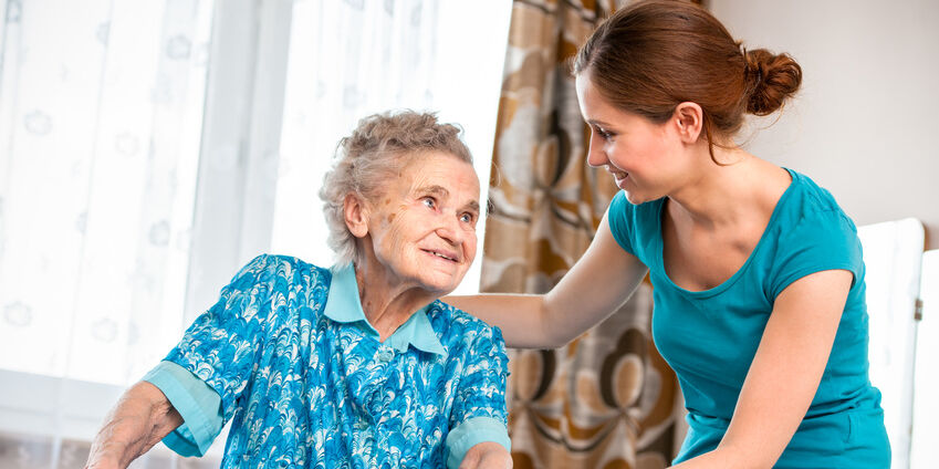Eine Seniorin geht an einer Gehhilfe und bekommt hierbei Unterstützung von einer jungen Pflegerin. Die beiden schauen sich an.
