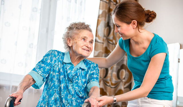 Eine Seniorin geht an einer Gehhilfe und bekommt hierbei Unterstützung von einer jungen Pflegerin. Die beiden schauen sich an.