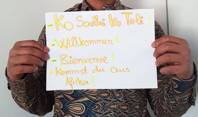 Ein farbiger Mann hält ein selbstgeschriebenes Schild hoch. Daraus steht "Willkommen" in verschiedenen Sprachen.