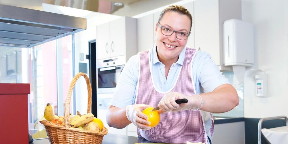 Eine Mitarbeiterin steht in einer Küche und schneidet eine Orange. Neben ihr steht ein Korb mit viel Obst ud vor ihr steht ein Teller auf welchem bereits aufgeschnittenes Obst liegt. 