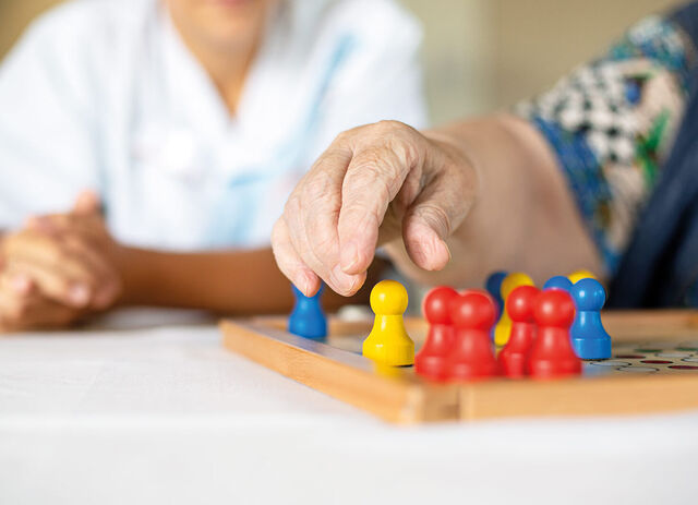 Ein Spielbrett für Menschärgeredichnicht. Eine Hand greift nach einer der Spielfiguren und zusätzlich befindet sich noch eine weitere Person am Tisch.
