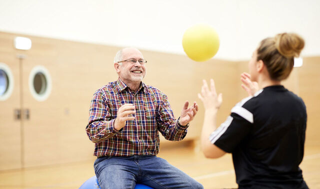 Eine Therapeutin wirft einem sitzenden älteren Patienten einen gelben Ball zu. Dieser fängt ihn gut gelaunt.