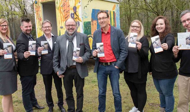 Team schaut in die lächelnd in die Kamera und jeder hält einen Flyer der Augustinus Gruppe in der Hand. und zeigt diesen in die Kamera.