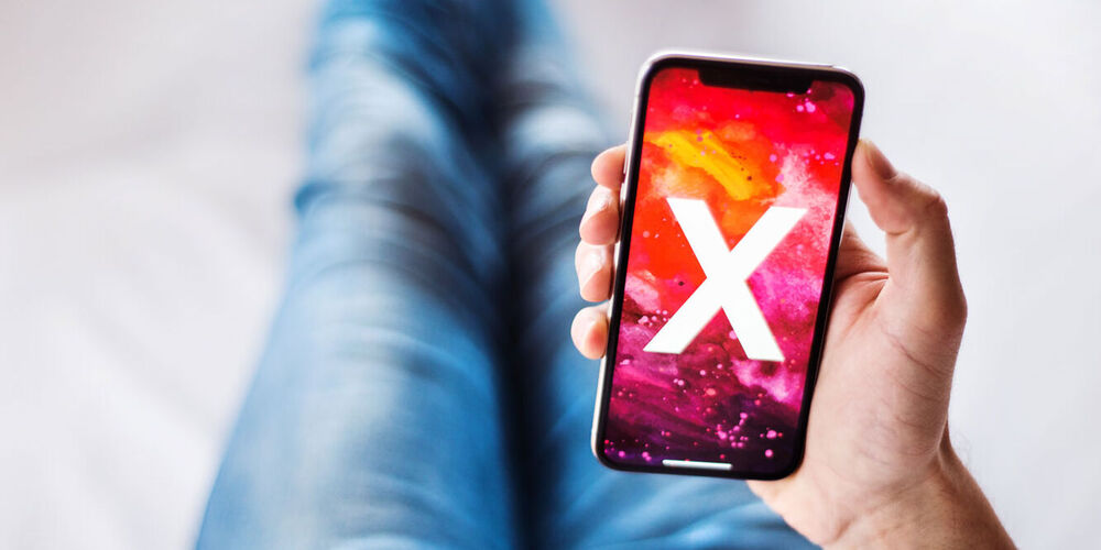 Ein Mann sitzt auf einem Boden und hält ein Smartphone in der Hand, auf welchem ein X ist. 