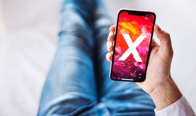 Ein Mann sitzt auf einem Boden und hält ein Smartphone in der Hand, auf welchem ein X ist. 