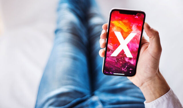Ein Mann sitzt auf einem Boden und hält ein Smartphone in der Hand, auf welchem ein X ist.