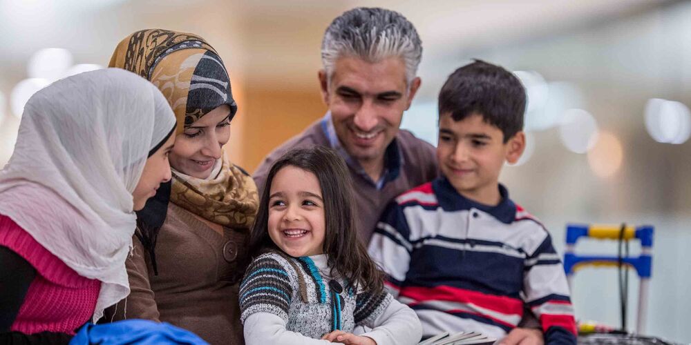 Eine Familie mit Flüchtlingshintergrund sitzt lächelnd beisammen. Sie besteht neben den Eltern aus drei Kindern.