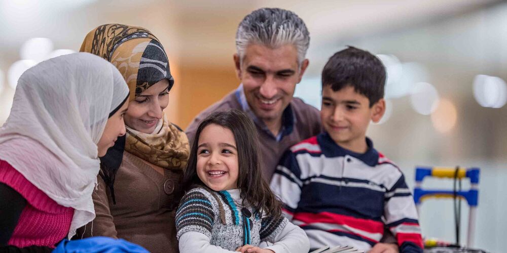 Eine Familie mit Flüchtlingshintergrund sitzt lächelnd beisammen. Sie besteht neben den Eltern aus drei Kindern. 