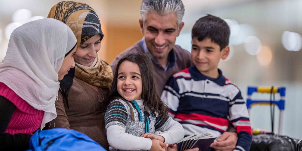 Eine Familie mit Flüchtlingshintergrund sitzt lächelnd beisammen. Sie besteht neben den Eltern aus drei Kindern. 