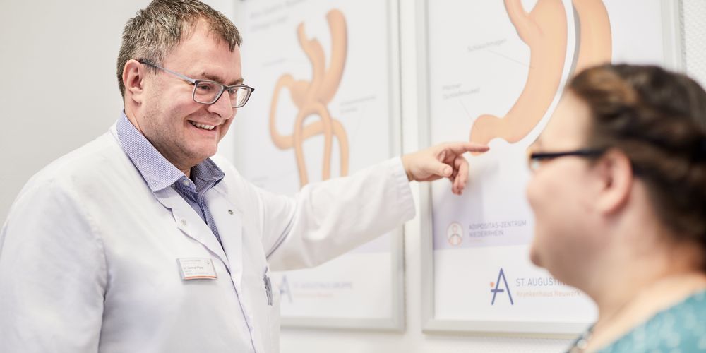 Ab wann sollte Adipositas behandelt werden? Ein Arzt erklärt einer Patientin anhand eines Schaubildes des Magens, warum eine Adipositas-Behandlung wichtig ist.
