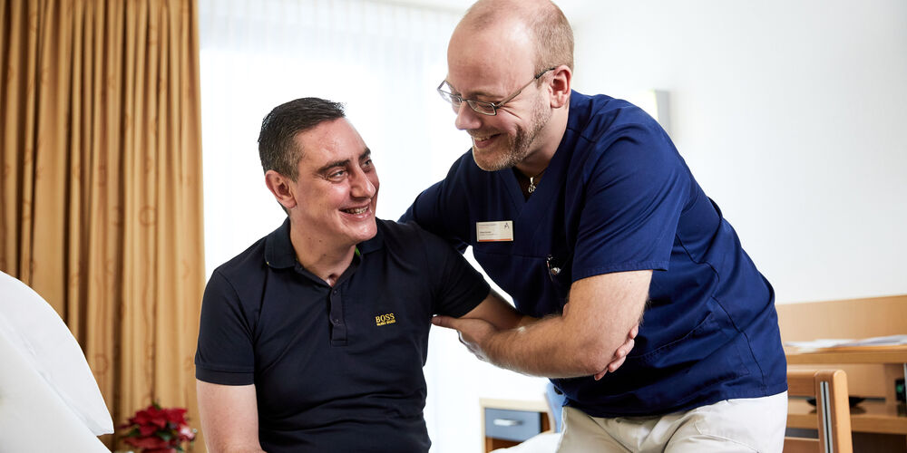 Ein Mitarbeiter der Reha hilft einem Patienten beim Aufstehen 