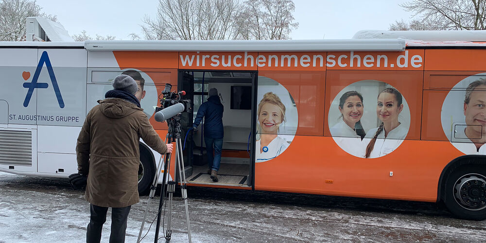Eine Straße auf der ein Mann mit einer Kamera steht und filmt wie ein weiterer Mann in einem Bus steigt, der Werbung von die Augustinus Gruppe trägt.