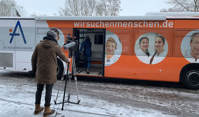 Eine Straße auf der ein Mann mit einer Kamera steht und filmt wie ein weiterer Mann in einem Bus steigt, der Werbung von die Augustinus Gruppe trägt.