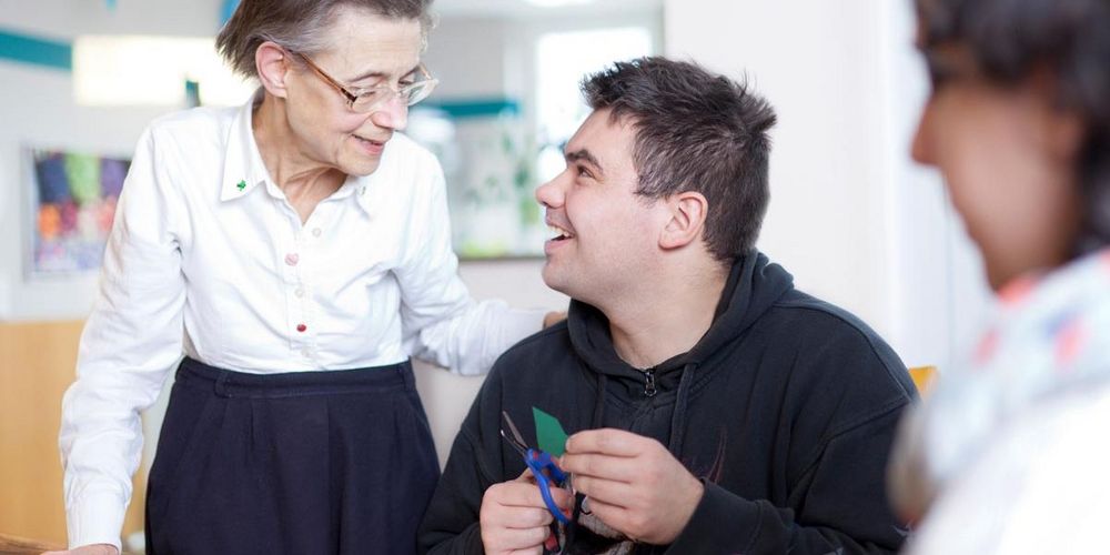 Eine ältere Dame steht neben einem jungen Mann, welcher an einem Tisch sitzt und bastelt. Sie hat ihm eine Hand auf die Schulter gelegt. Eine weitere jüngere Frau sitzt mit am Tisch. 