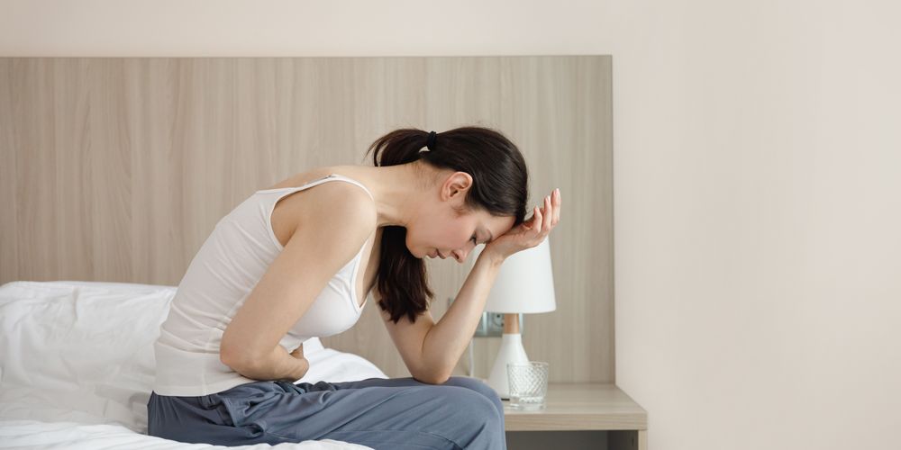 Woran machen sich Dranginkontinenz Symptome bemerkbar? Eine Frau sitzt auf einem Bett und leidet unter Dranginkontinenz Symptomen.